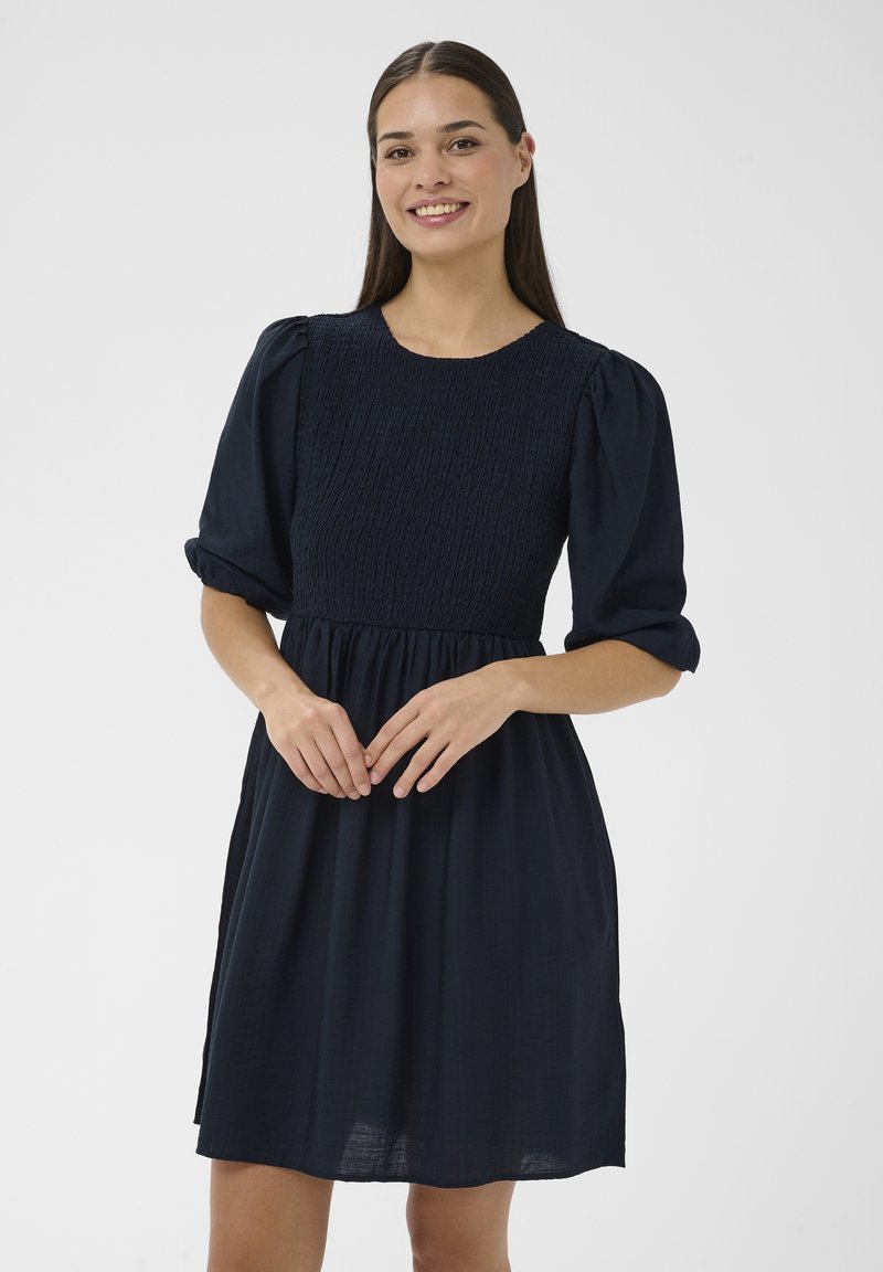 Femme souriante aux longs cheveux bruns, portant une robe bleu marine à manches bouffantes et corsage smocké, debout devant un fond uni.