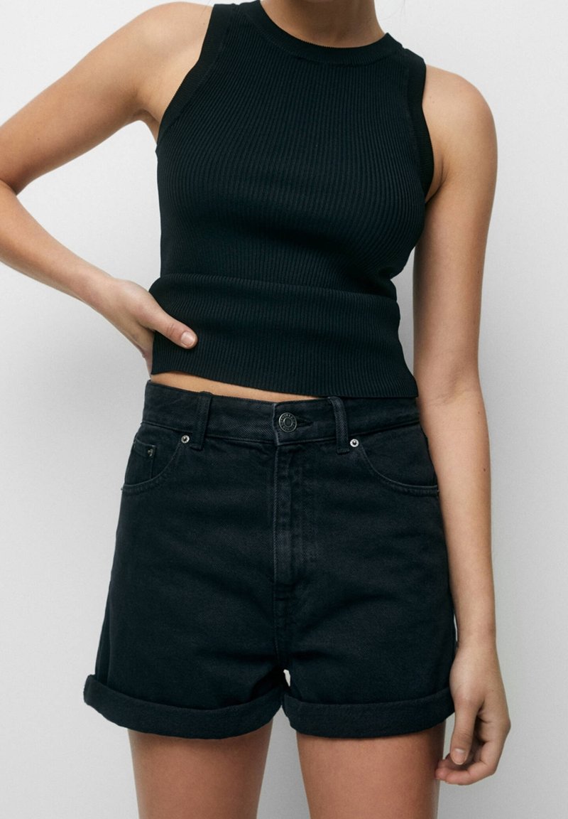 Personne portant un crop top noir côtelé sans manches et un short en denim noir à ourlet retroussé, debout avec une main sur la hanche devant un fond uni.