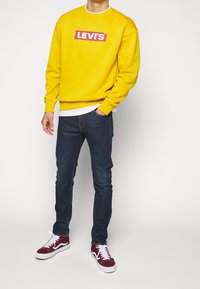 Sudadera amarilla con cuello redondo blanco y logo rojo de "LEVI'S", combinada con vaqueros azul oscuro y zapatillas burdeos con suelas blancas.