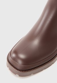 Gros plan sur une botte en caoutchouc marron avec un embout rond et une semelle texturée sur fond blanc.