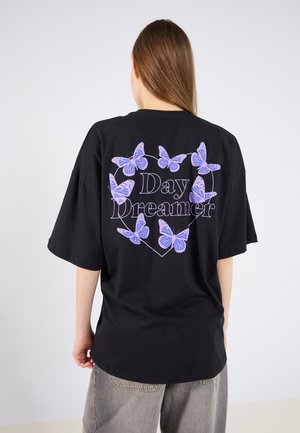 Persoon met lang haar draagt een zwart oversized T-shirt met paarse vlinders en de tekst "Day Dreamer" binnen een hart op de rug.