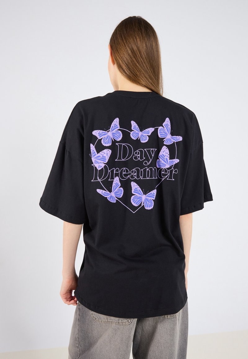 Personne aux longs cheveux portant un T-shirt noir oversize avec des papillons violets et le texte "Day Dreamer" à l'intérieur d'un cœur dans le dos.
