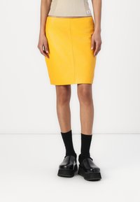 Geel leren kokerrok met een aansluitend ontwerp, voorzien van een gladde textuur. Gepaard met zwarte plateau schoenen en sokken.