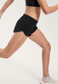 Čierne športové šortky s voľným strihom a bočnými rozparky, vyrobené z ľahkého materiálu, v kombinácii s neutrálne zafarbenými bežeckými topánkami.