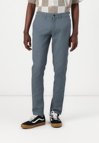 Grå bomullschinos med slim fit, med platt front och sidfickor, i kombination med svarta sneakers och en rutiga skjorta med knappar.