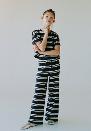 Jeune personne portant un haut à manches courtes à rayures noires et blanches et un pantalon large assorti, debout avec la main posée sur le menton devant un fond uni.