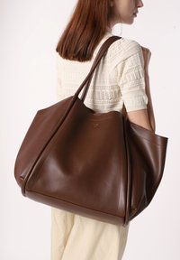 Bolso tote de cuero marrón con textura suave, forma grande y asas delgadas, que presenta costuras mínimas y una sutil marca en la parte frontal.