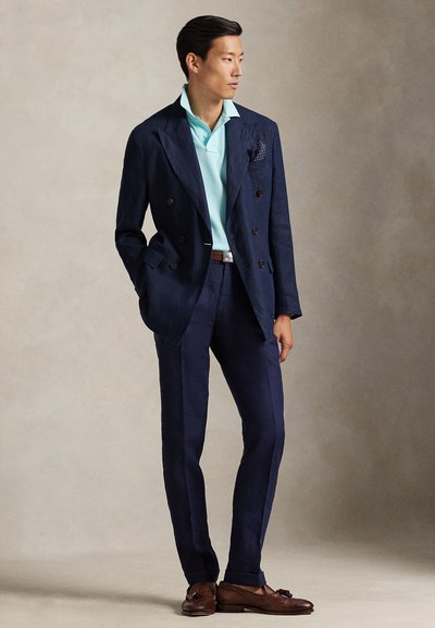 Δίπτυχος σακάκι από λινό σε ναυτικό μπλε, συνδυασμένο με ανοιχτό μπλε πόλο πουκάμισο, ναυτικο μπλε παντελόνι και καφέ μοκασίνια. Μαντηλάκι τσέπης προσθέτει λεπτομέρεια.