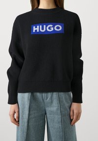 Schwarzer Strickpullover mit geripptem Muster, versehen mit einem blauen rechteckigen Logo mit "HUGO" in weißen Buchstaben über der Brust.