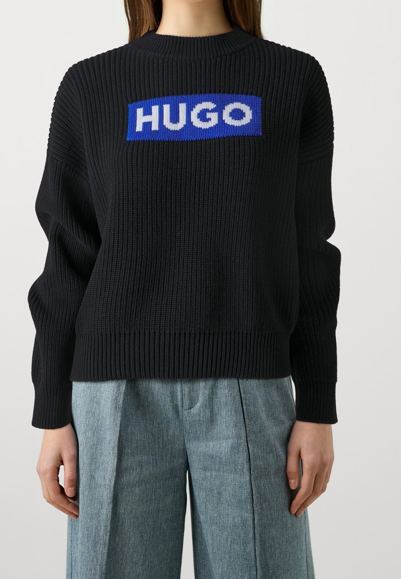 Schwarzer Strickpullover mit geripptem Muster, versehen mit einem blauen rechteckigen Logo mit "HUGO" in weißen Buchstaben über der Brust.