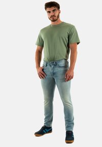 Uomo in una t-shirt a maniche corte verde chiaro e jeans slim-fit blu chiaro, che indossa sneakers blu scuro con strisce blu chiaro.