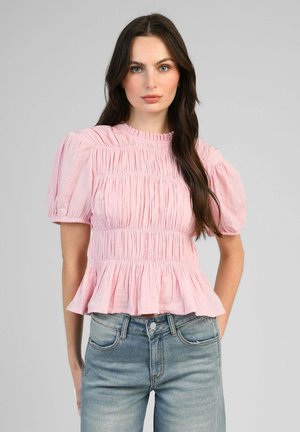 Femme aux longs cheveux foncés porte un chemisier rose froncé avec manches bouffantes et ourlet péplum, assorti à un jean taille haute délavé clair.