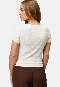 Short-sleeve top in cream textured fabric with a quilted pattern, featuring a round neckline and fitted style, paired with brown pants.