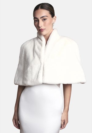 Donna con capelli pettinati all'indietro che indossa un morbido mantello di pelliccia bianca sopra un abito bianco senza maniche su uno sfondo chiaro e semplice.