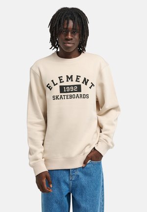 Młoda osoba z ciemnymi dredami, ubrana w beżową bluzę z napisem "ELEMENT 1992 SKATEBOARDS" i niebieskie dżinsy na jednolitym tle.