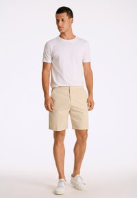 Shorts beiges avec une taille élastique, t-shirt blanc à manches courtes et baskets blanches. Le modèle se tient contre un fond uni.