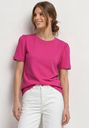 Ung kvinde med brunt hår, der er sat tilbage, iført en lys pink kortærmet top og hvide bukser, smilende og kiggede til siden.