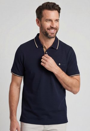 Man die lacht, een marineblauw poloshirt met beige bies draagt, zijn kraag aanraakt, staand tegen een effen lichte achtergrond.