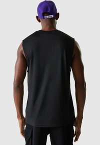 Schwarzes ärmelloses Sportshirt aus glattem Stoff, mit rundem Ausschnitt und geradem Saum. Getragen mit einer lila Baseballkappe.