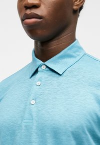 Νεαρό άτομο με σκουρόχρωμο δέρμα που φοράει ένα ανοιχτό μπλε πόλο με κουμπιά, ορατό από τον λαιμό μέχρι το πάνω μέρος του στήθους, σε φόντο μονόχρωμο.