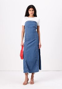 Denim maksi obleka s strukturiranim korpusom, belimi T-majicami in vidnim šivanjem; model drži rdečo torbico in nosi sandale na vezalke.