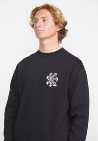 Sudadera negra con un diseño gráfico blanco en el área del pecho izquierdo, con un clásico cuello redondo y puños de canalé. La tela parece suave y texturizada.