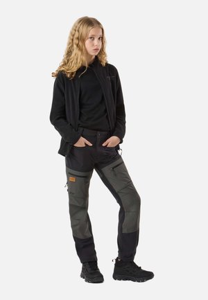 Giovane donna con capelli biondi e ricci che indossa una giacca nera, pantaloni cargo neri e grigi e scarponi da trekking neri, in piedi con le mani in tasca.