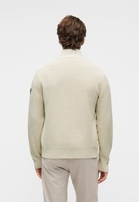 Φωτεινό εκρού πουλόβερ με υφή πλεκτού, ψηλό γιακά και ριμπ μανσέτες. Διαθέτει διακριτικό λογότυπο στο αριστερό μανίκι. Συνδυάζεται με μπεζ παντελόνια.