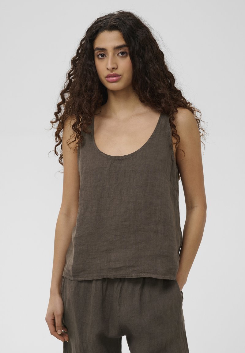 Jeune femme aux longs cheveux bouclés porte un haut marron ample sans manches et un pantalon assorti, debout devant un fond clair uni.
