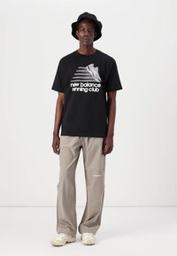 Juodas marškinėliai su balta "new balance running club" grafika, derinami su šviesiais smėlio spalvos plačiomis kelnėmis ir smėlio spalvos sportiniais bateliais. Apsaugantis juodas skrybėlė.