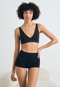 Schwarzer Bralette mit V-Ausschnitt und breiten Trägern, kombiniert mit hochgeschnittenen schwarzen Shorts. Beide bestehen aus weichem, dehnbarem Stoff.