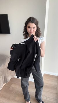 Jeune femme aux cheveux bouclés tenant un t-shirt noir à manches longues dans une pièce peu meublée avec des murs clairs et un sol en bois.