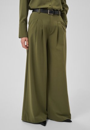 Persoon met olijfgroene, wijde pantalon met plooien, een zwarte riem en bijpassend overhemd; alleen het onderlichaam is zichtbaar.