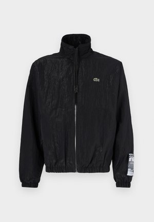 Black lightweight jacket made from textured fabric, featuring a high collar, zip front, and elastic cuffs with a logo patch on the sleeve.