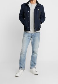 Marine bomberjack met zwarte accenten, ritszakken en een logo patch. Gecombineerd met een lichtgrijs t-shirt, blauwe jeans en witte sneakers.