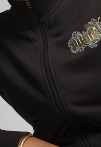 Chaqueta negra con cremallera hecha de un tejido suave, con un diseño floral dorado y un logo en la parte superior izquierda. Detalle de cremallera visible.