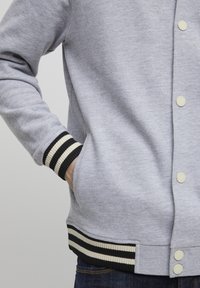 Grå hettegenser med frontknapper, ribbede svarte og hvite mansjetter, og sidelommer. Glatt tekstur med et uformelt design.