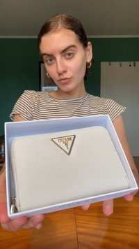 White leather wallet presented in a white box, featuring a gold triangular logo accent. The wallet has a textured surface and zip closure.