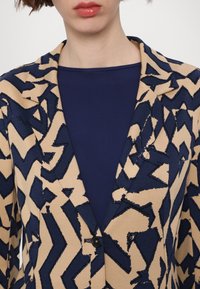 Femme portant un blazer beige avec un motif abstrait bleu marine audacieux sur un haut bleu marine uni, lèvres avec un rouge à lèvres brillant rouge visible.