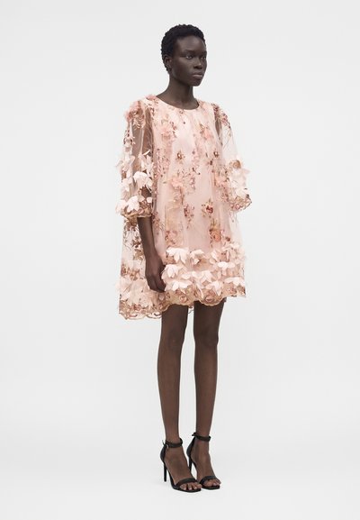 Γυναίκα που στέκεται φορόντας ένα κοντό ροζ διάφανο φόρεμα με floral εφαρμογές, φουσκωτά μανίκια και μαύρα ψηλοτάκουνα σανδάλια σε απλό φόντο.
