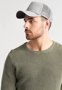 Casquette de baseball grise avec un devant texturé, des panneaux latéraux en maille et une visière droite, associée à un pull en laine vert olive.