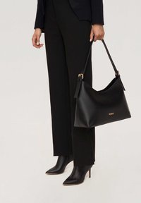 Schwarze Lederhandtasche mit glatter Textur, geschwungener Form und goldenem Logo. Kombiniert mit eng anliegenden schwarzen Hosen und spitzen schwarzen Stiefeln mit Absatz.