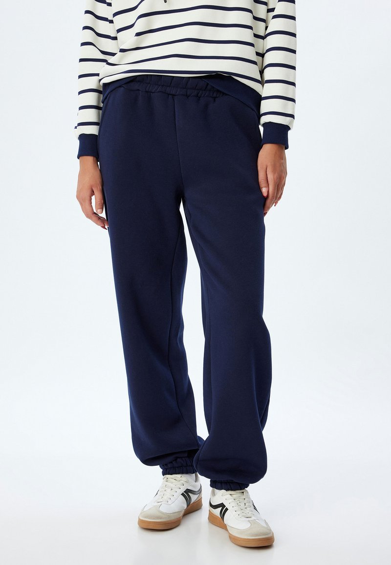 Marineblå sweatpants med elastisk talje og manchetter ved anklerne, lavet af blødt stof. Kombineret med hvide sneakers med sorte detaljer.