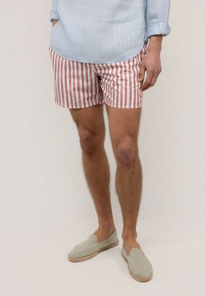 Hombre con camisa de manga larga azul claro remangada, pantalones cortos de rayas rojas y blancas y zapatos beige sin cordones, de pie con las manos en los bolsillos.