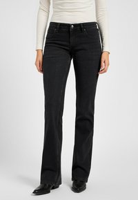 Frau in enganliegenden schwarzen Bootcut-Jeans, weißem gerippten Langarmoberteil und schwarzen spitzen Boots, die vor einem schlichten Hintergrund steht.