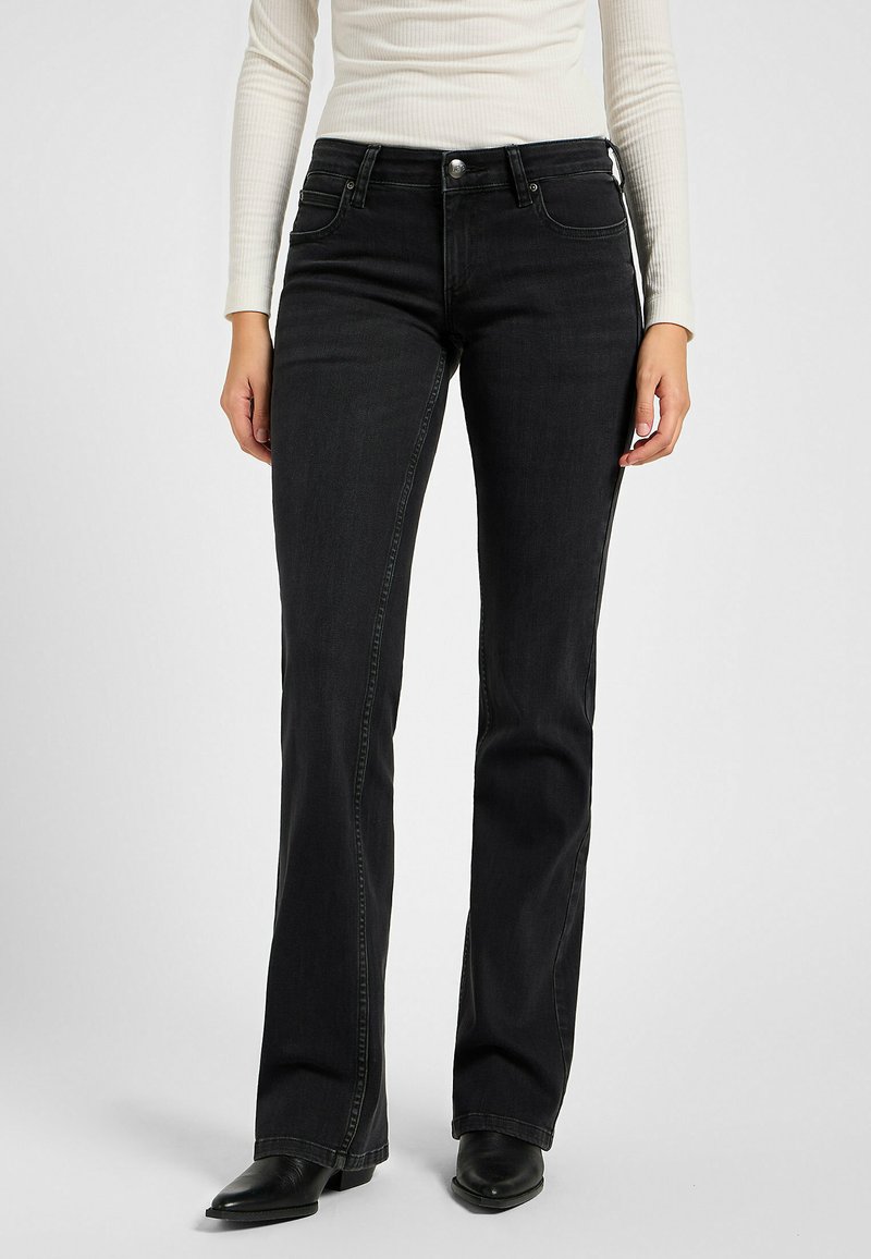 Frau in enganliegenden schwarzen Bootcut-Jeans, weißem gerippten Langarmoberteil und schwarzen spitzen Boots, die vor einem schlichten Hintergrund steht.