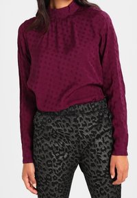 Blouse bordeaux à manches longues avec une texture subtile à pois et un col montant, associée à un pantalon noir ajusté orné d'un imprimé léopard métallique.