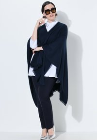 Marineponcho mit Fledermausärmeln und Fransen, darüber ein weißes Hemd und schwarze Hosen, kombiniert mit silbernen Schuhen und übergroßen Sonnenbrillen.