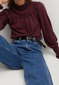 Bordeaux top met een hoge gerimpelde hals, lange mouwen en gather-schouders, gecombineerd met lichtblauwe hoge taille jeans en een zwarte riem.