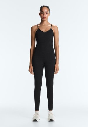 Žena stoji i nosi crni uski kombinezon bez rukava s tankim naramenicama i bijele tenisice na običnoj svijetloj pozadini.